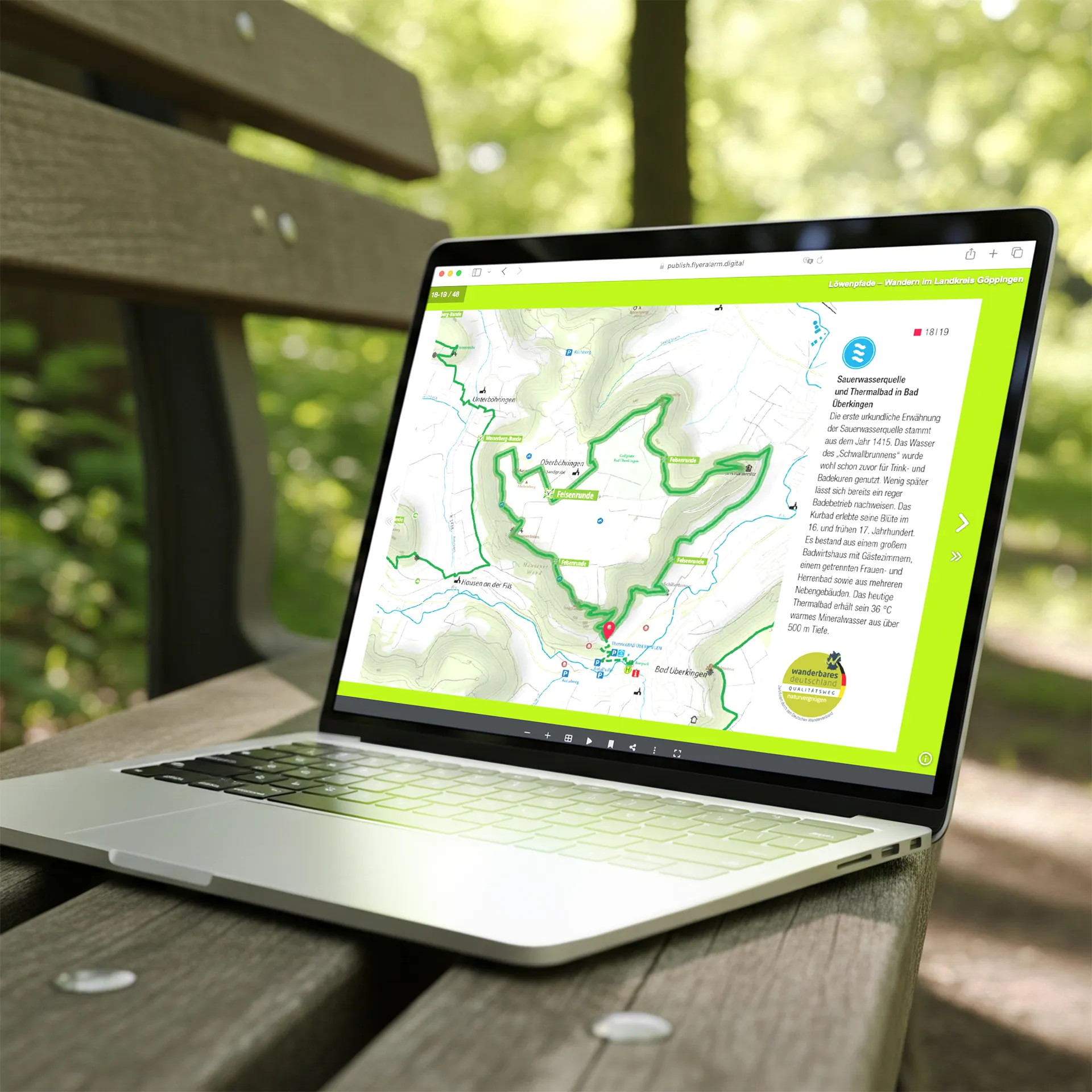 Digitale Löwenpfad-Wanderkarte auf Laptop Laptop auf einer Holzbank im Freien mit geöffnetem Fenster, das eine digitale Wanderkarte des Löwenpfad-Projekts im Landkreis Göppingen zeigt. Die Karte markiert Wanderrouten und Points of Interest wie die Sauerwasserquelle und das Thermalbad in Bad Überkingen. Im Hintergrund grüne Blätter und Lichtreflexe.